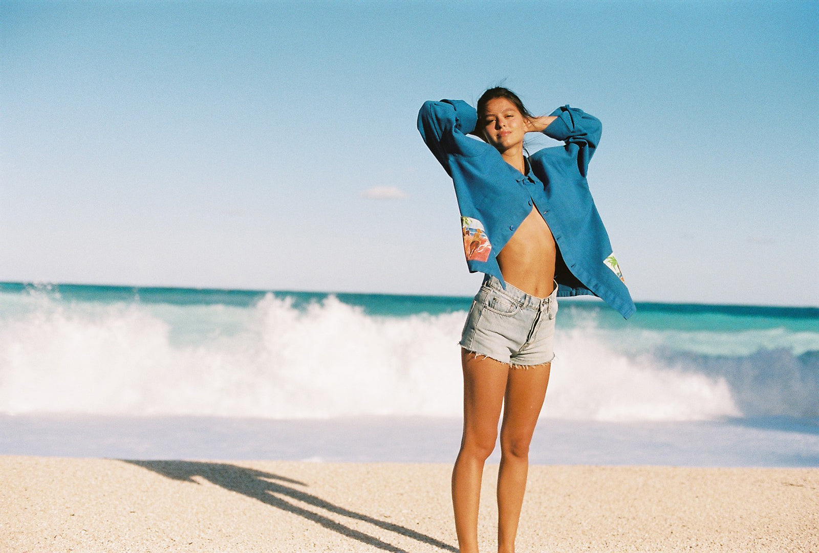 Femme en veste bleue et short en jean, posant sur une plage devant la mer